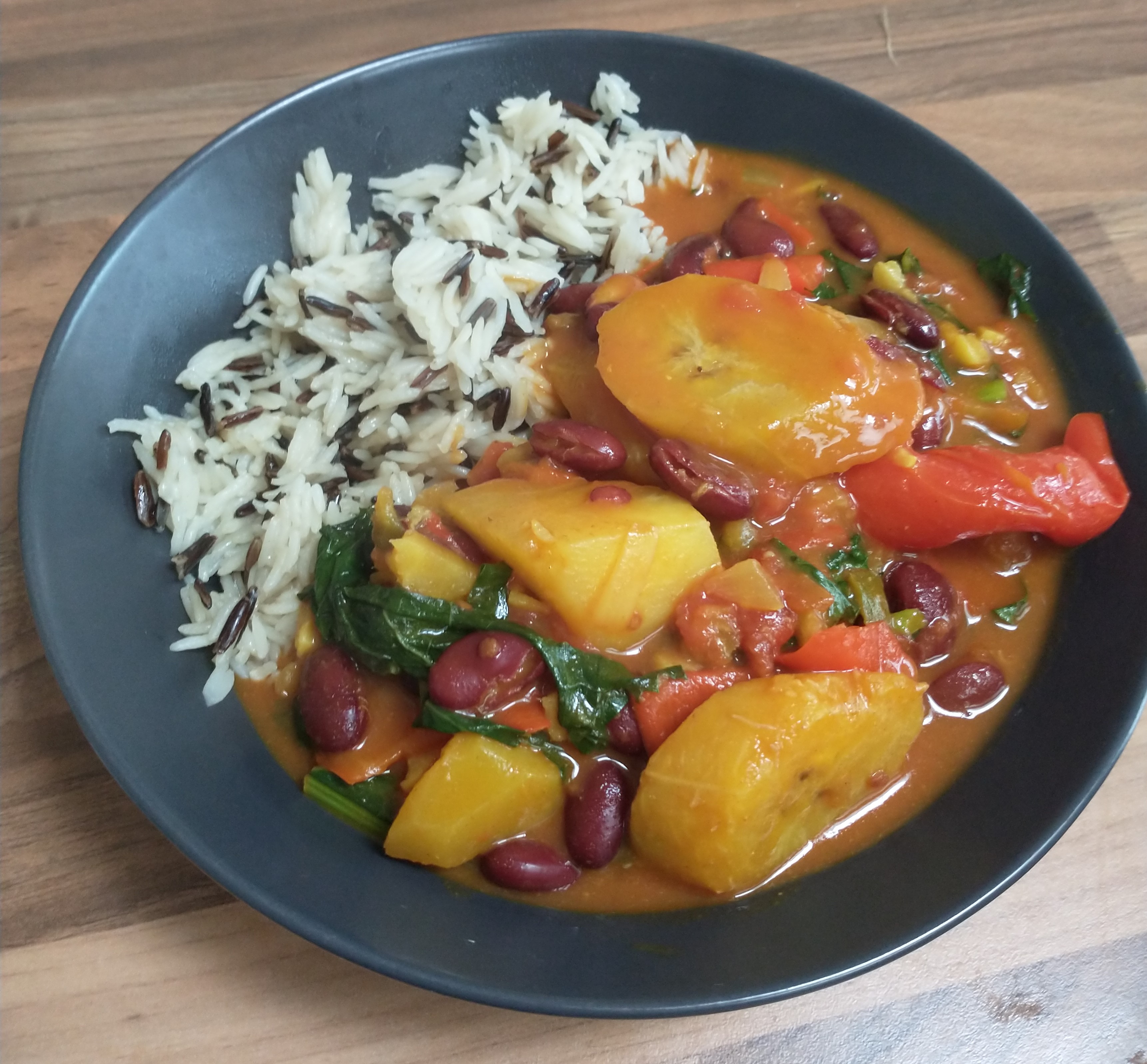 A picture of a flat bowl with wild rice, and a warm orange curry with visible plantain, peppers, spinach and kidney beans.